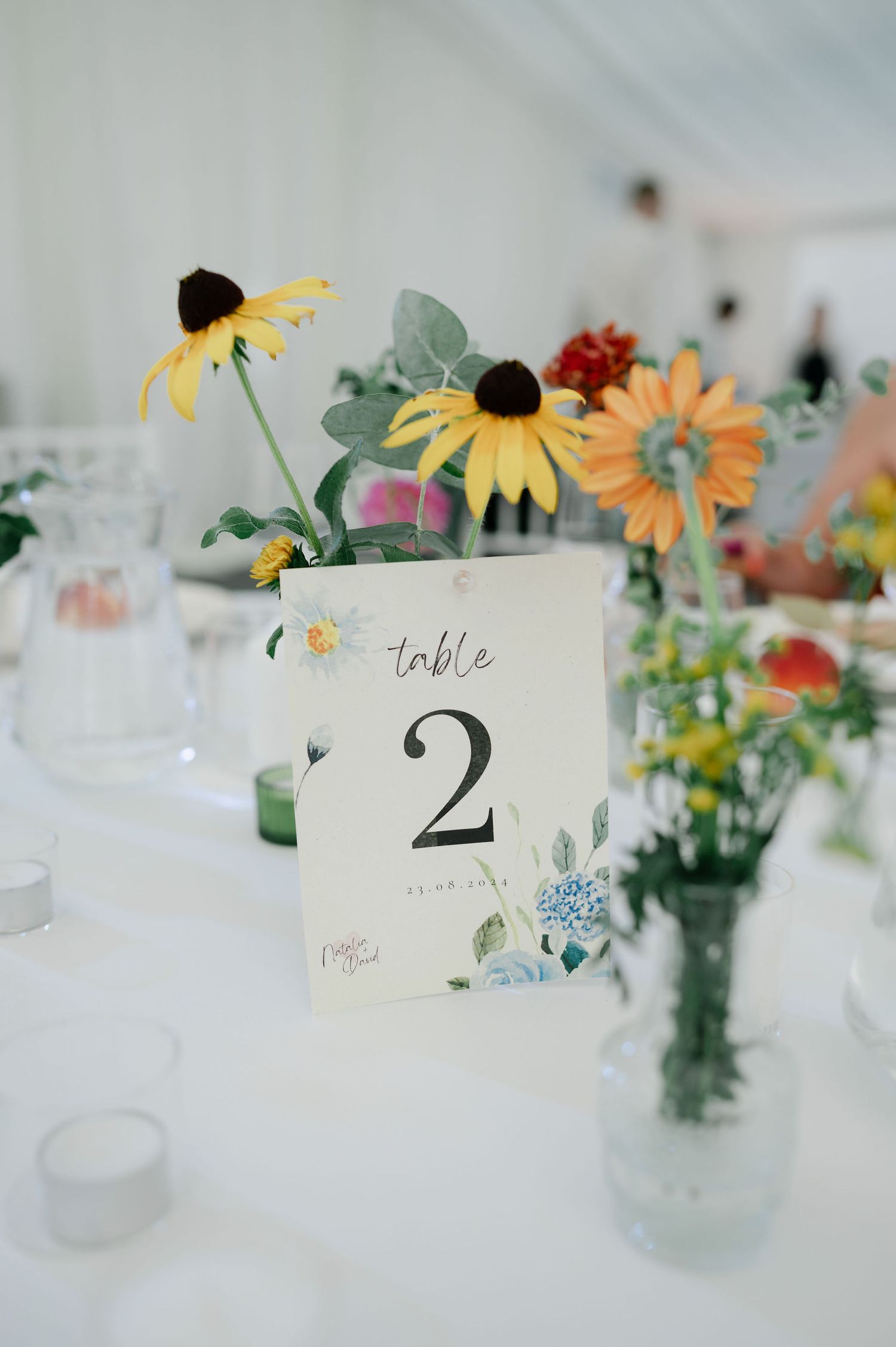 Wedding reception table number card surrounded by fresh summer flowers in glass vases.