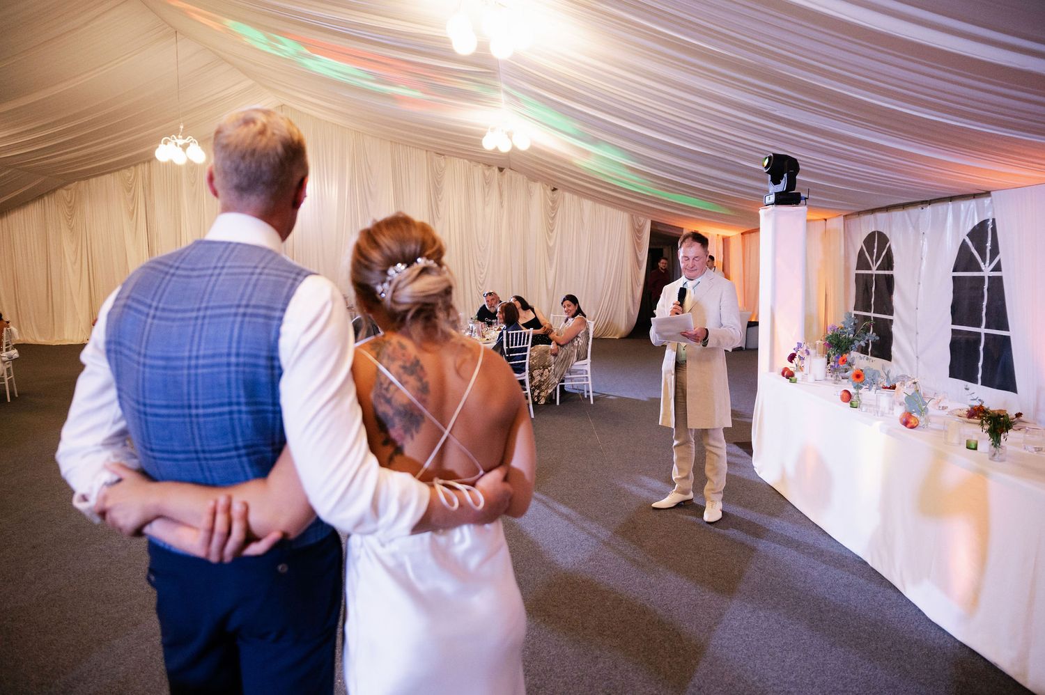 A couple listens to wedding speeches in an elegant tented reception space.