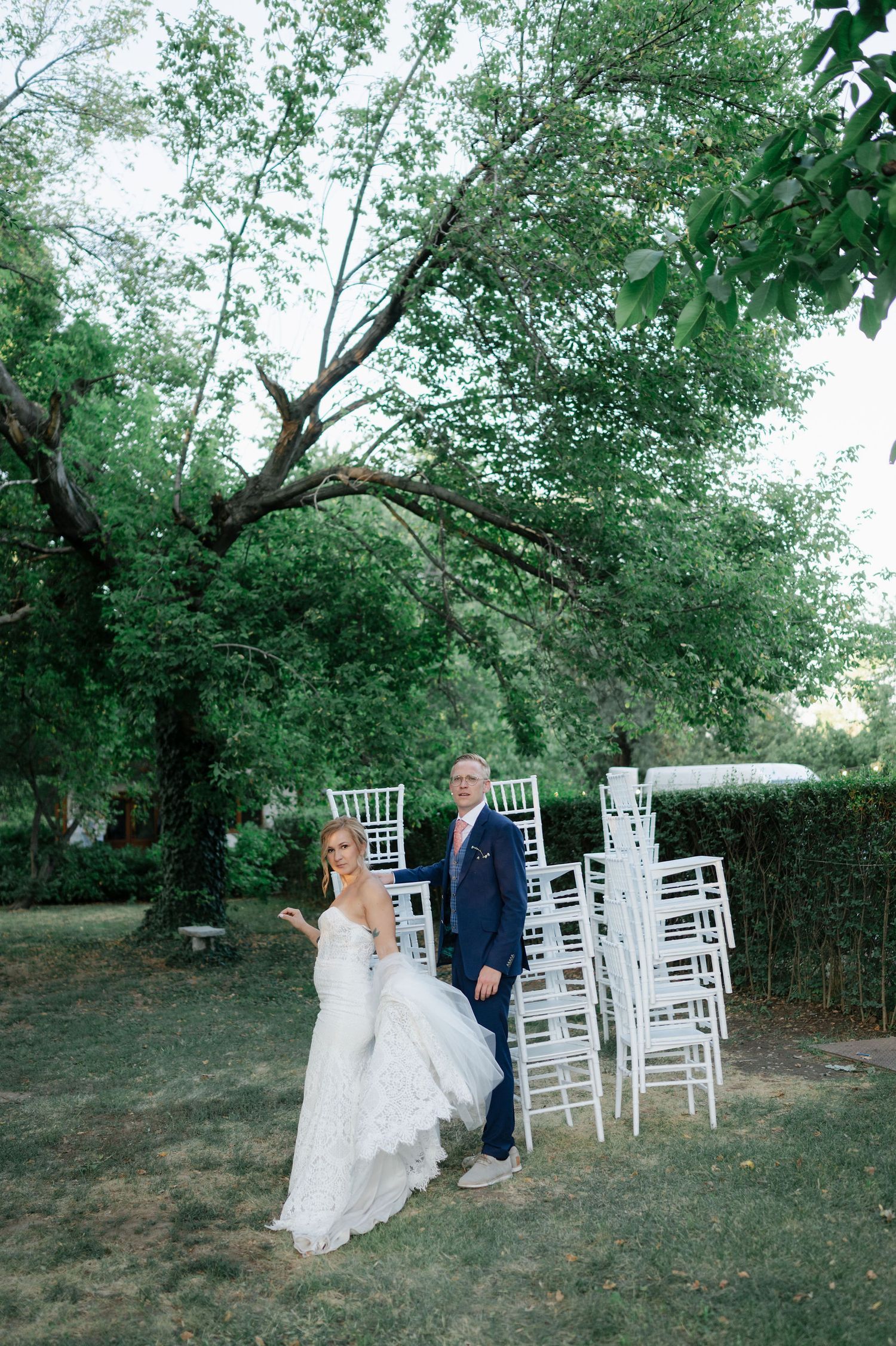 A couple poses near white wedding chairs arranged under leafy trees.