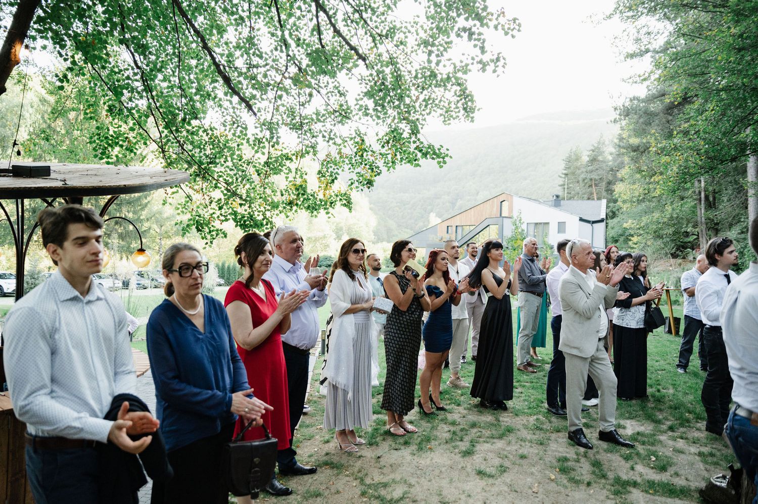 Wedding guests gather in a scenic wooded outdoor ceremony space with natural stone steps and greenery.