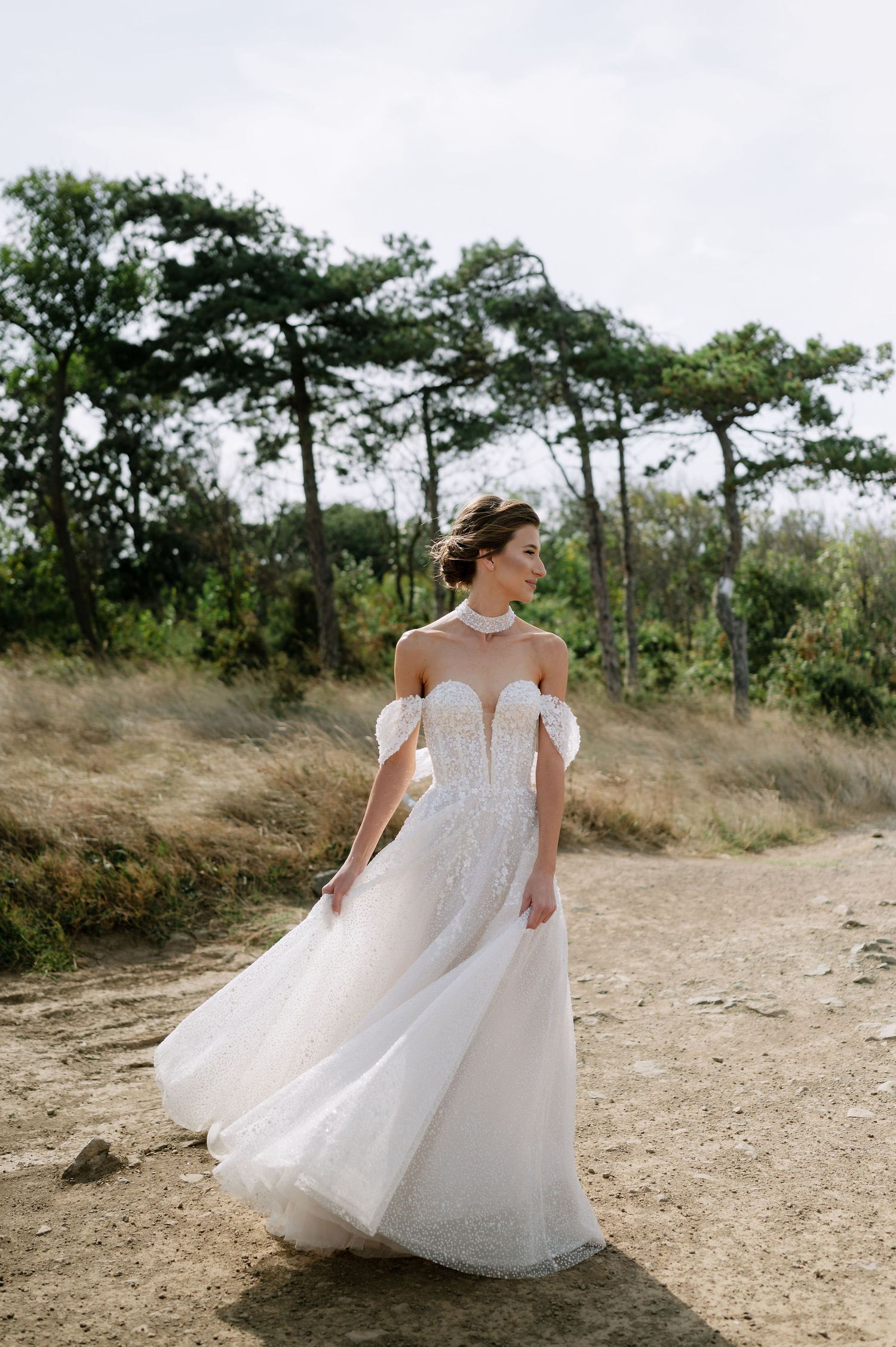 Bride in off-shoulder wedding dress poses gracefully on dirt path with trees in background.