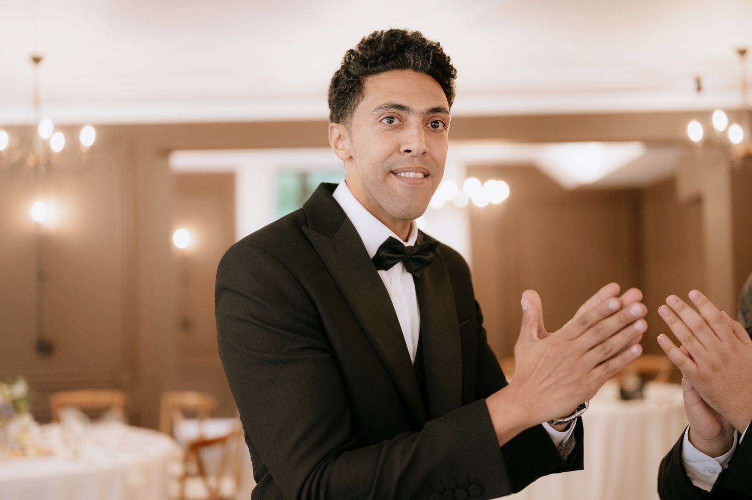 A groom in a black tuxedo smiling and gesturing during a wedding reception.