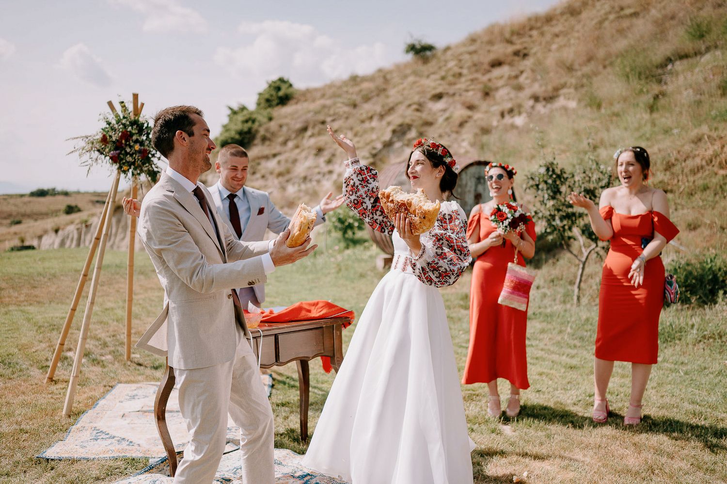 Outdoor wedding ceremony setup with red bridesmaid dresses and rustic decor against a hillside backdrop.