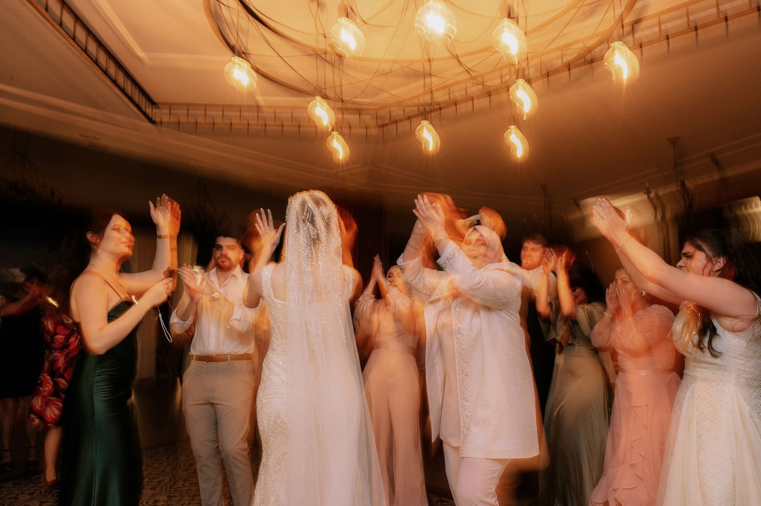 Wedding party dancing under warm string lights at an evening reception.