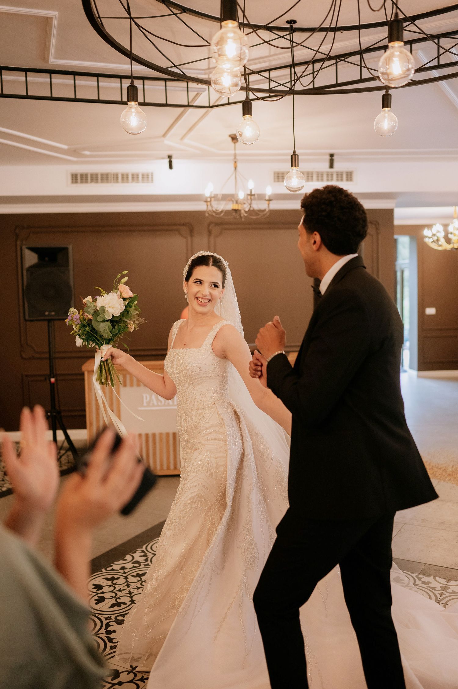 Wedding party members dance and celebrate in an indoor venue with hanging lights and decorative flooring.