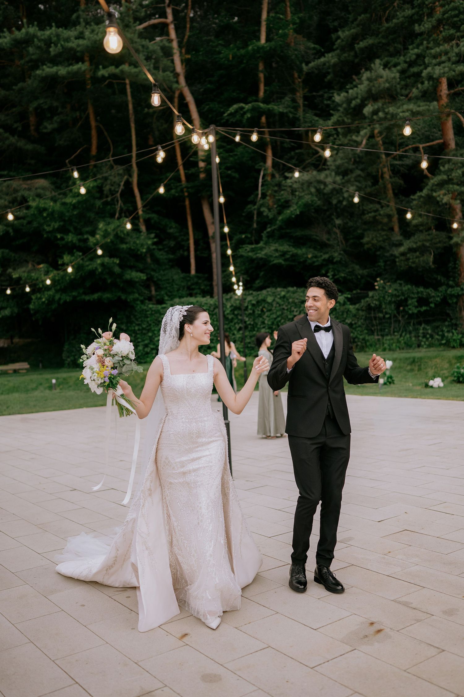 A couple dancing on an outdoor patio with string lights and trees in the background.