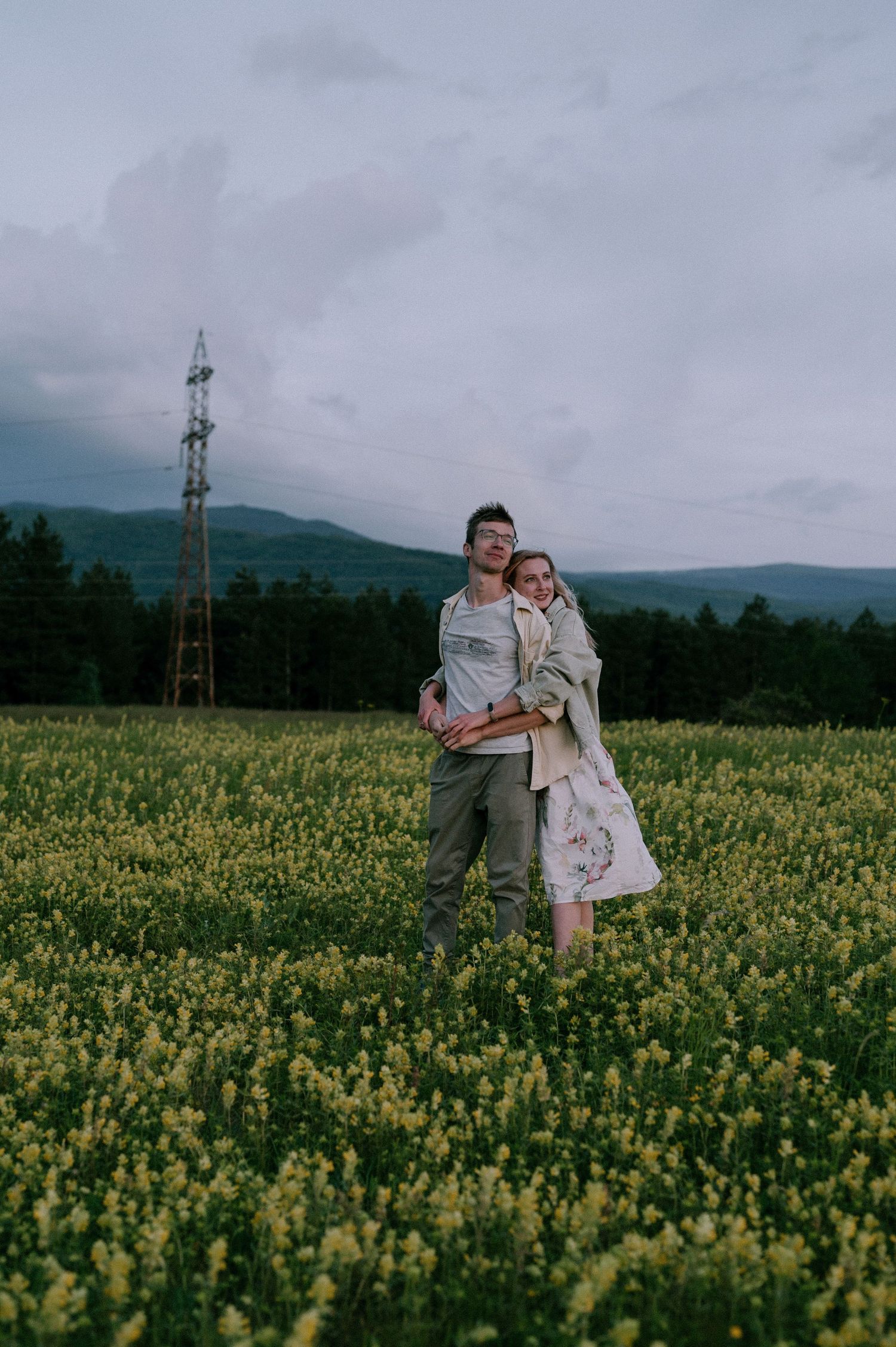 A couple shares an intimate moment in a field of yellow flowers with power lines in the distance.