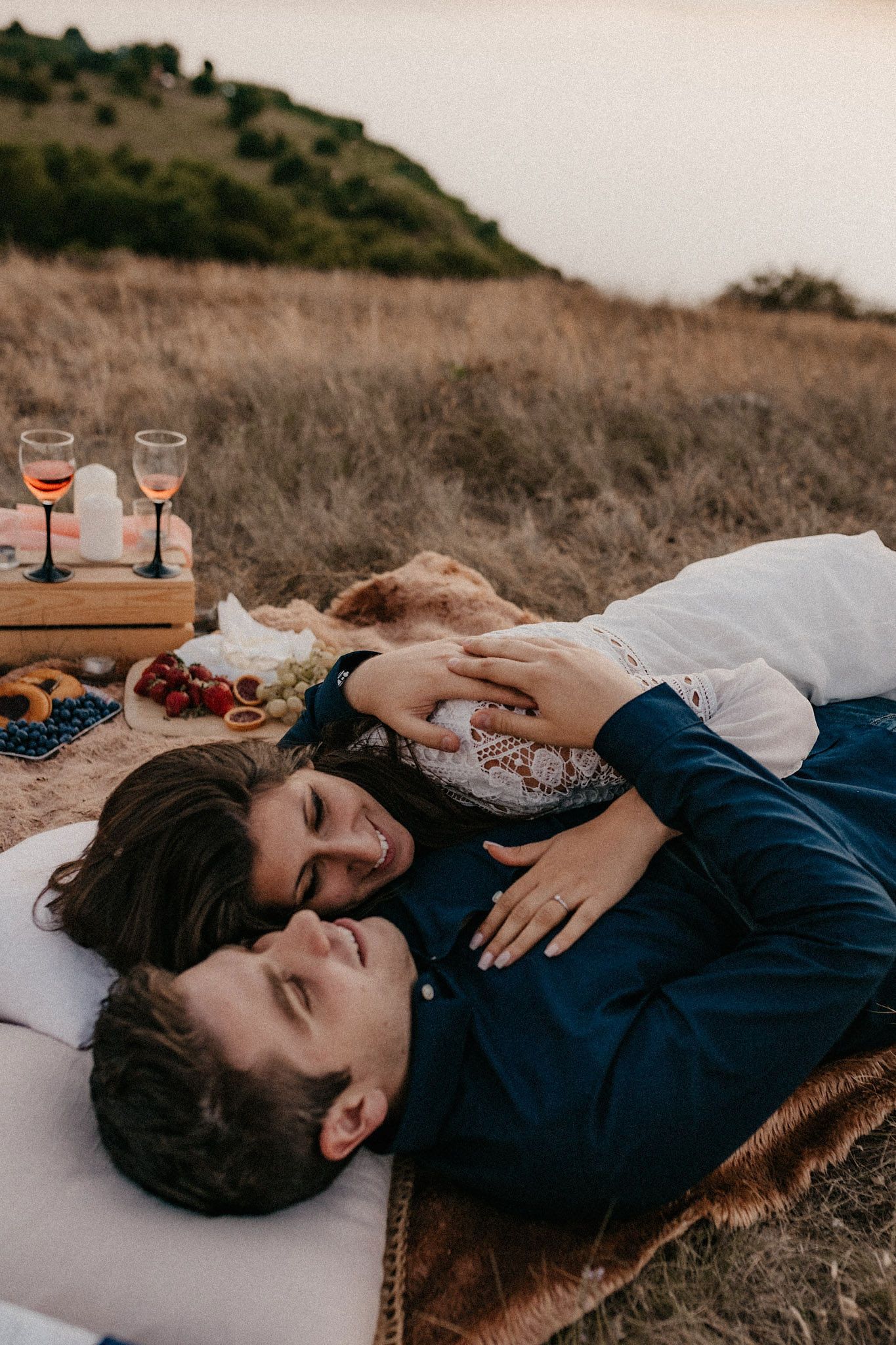 Couple relaxing on blanket during sunset picnic with wine and snacks.