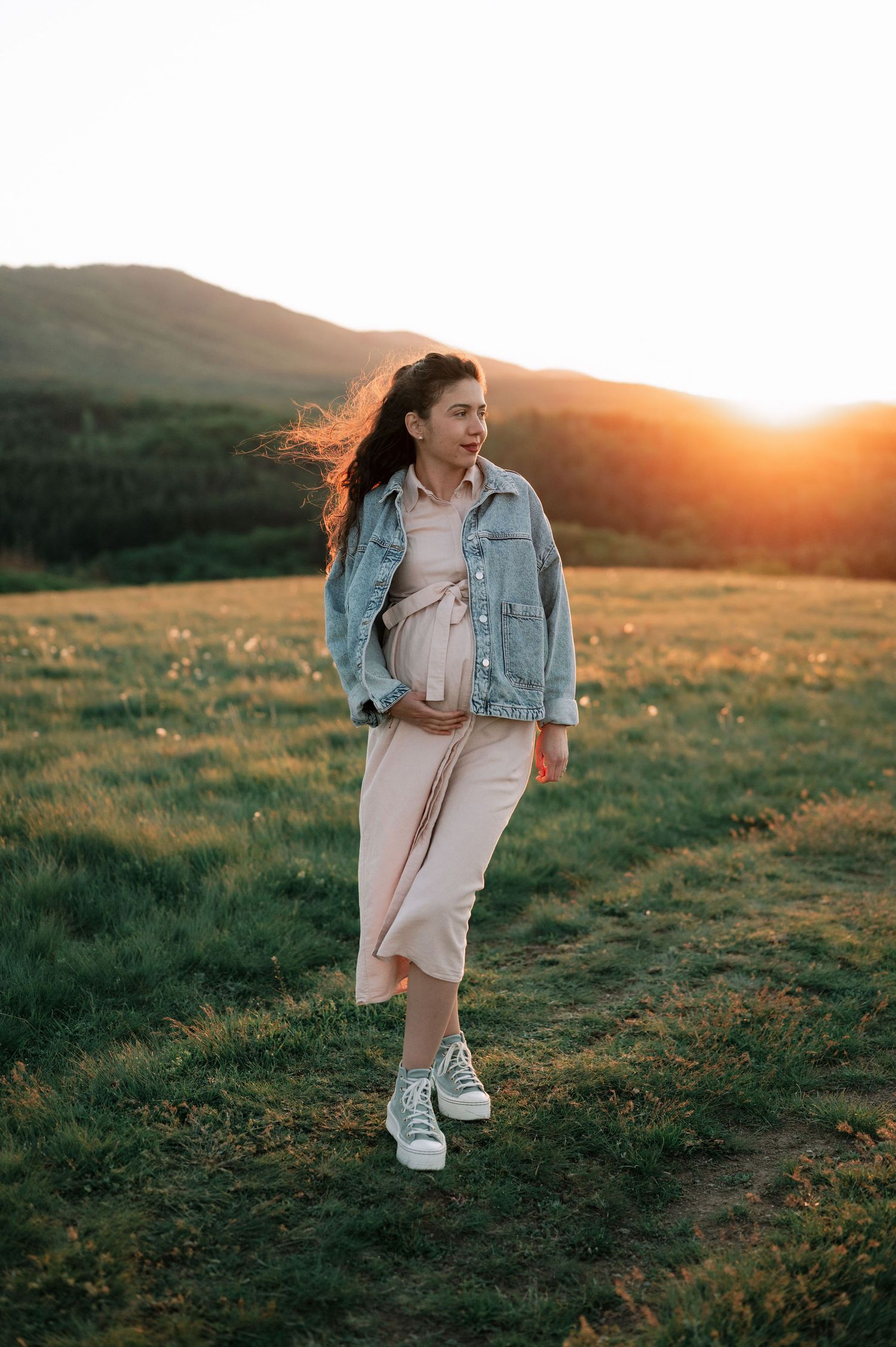 Person in a denim jacket and dress walks across a grassy hillside during golden hour sunset.