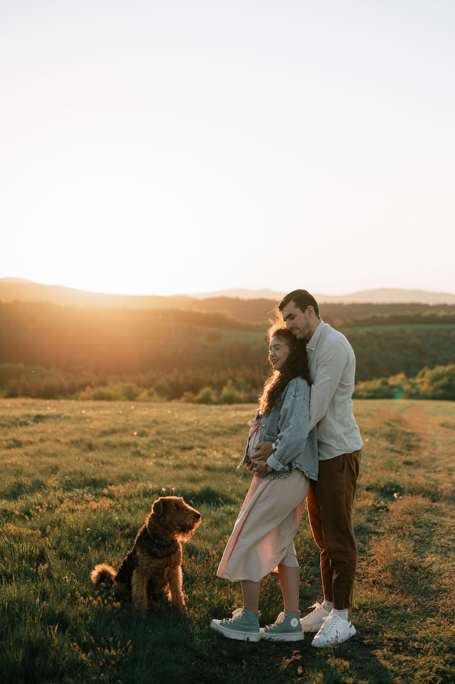 A couple and their dog enjoy a romantic sunset moment on a hilltop with golden light illuminating the landscape.