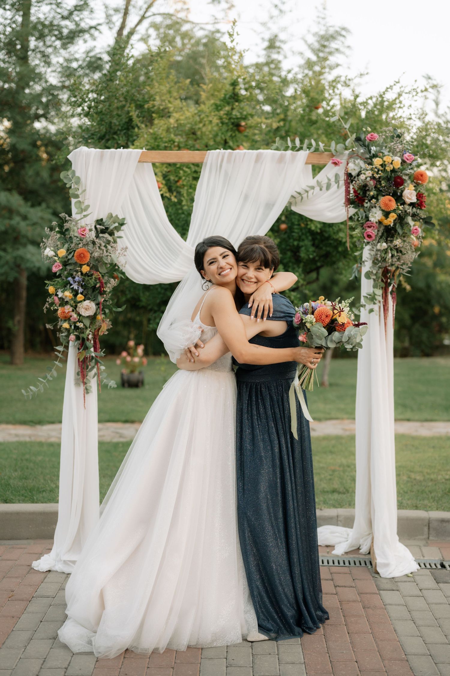 A beautiful outdoor wedding arch with white flowing fabric and coral floral arrangements on a brick patio.