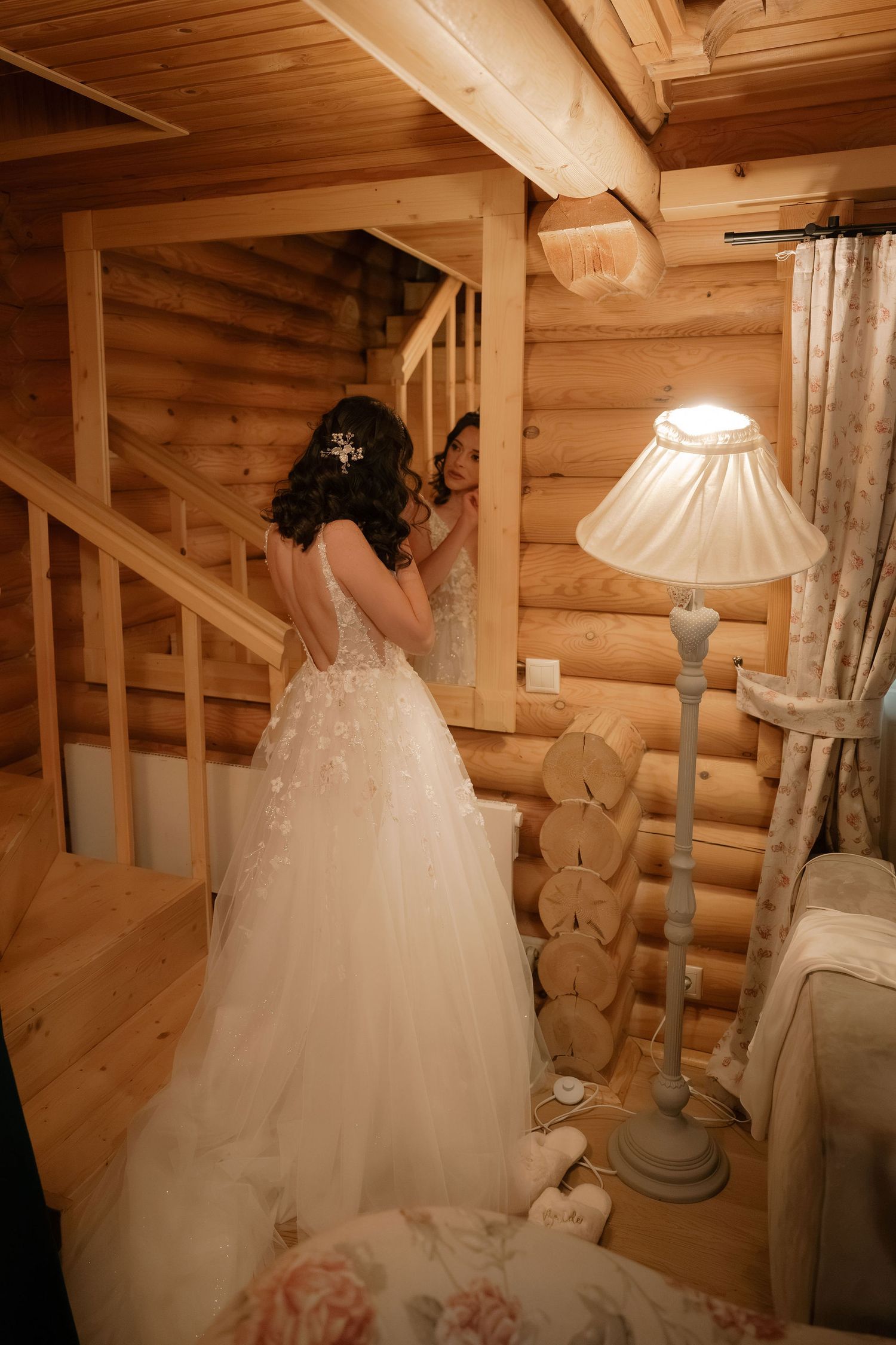Bride in elegant backless lace wedding gown standing by wooden log cabin staircase with vintage floor lamp.