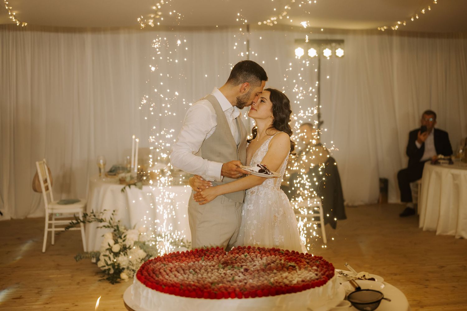Newlyweds share a kiss while cutting their wedding cake surrounded by sparkling fireworks at a romantic reception.