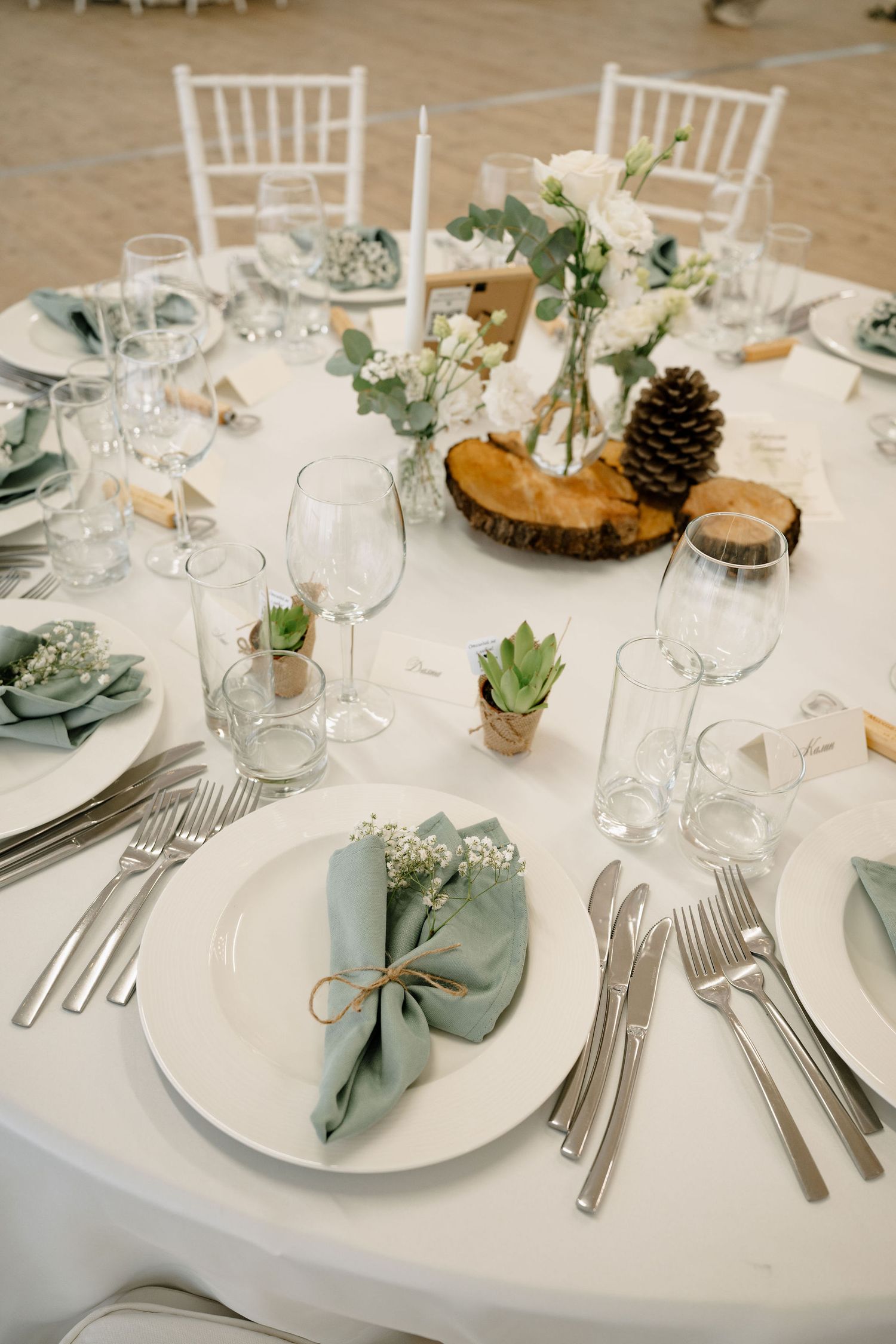 Elegant wedding table setting with sage green napkins, succulents, pine cone centerpiece, and white floral arrangements.