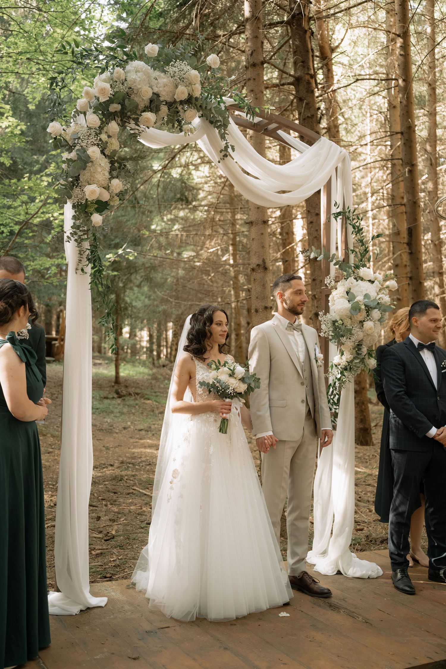 Bride and groom stand under floral arch with white draping in forest wedding ceremony.