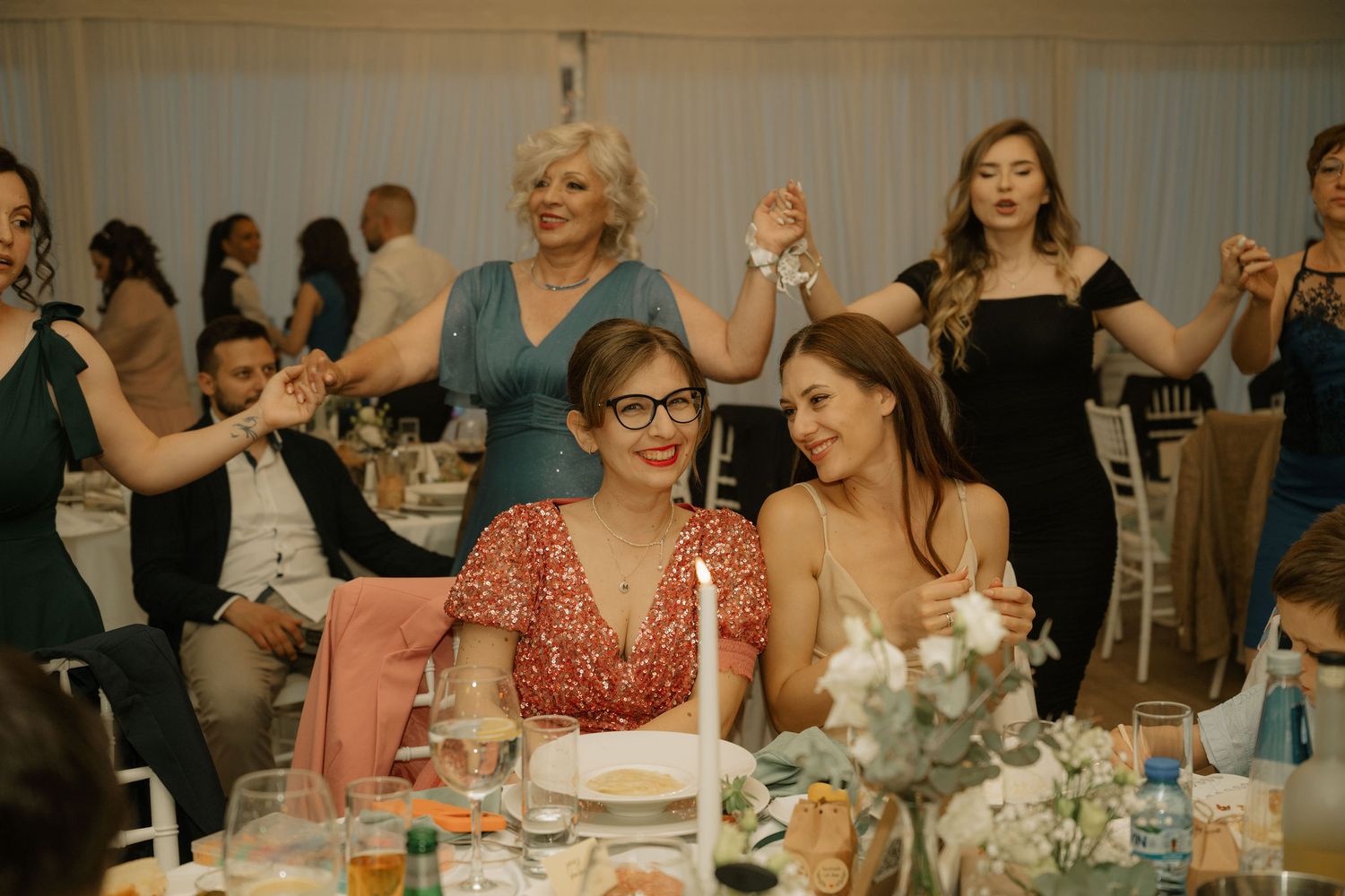 Two women smile at a wedding reception table while guests dance and celebrate in an elegant venue.