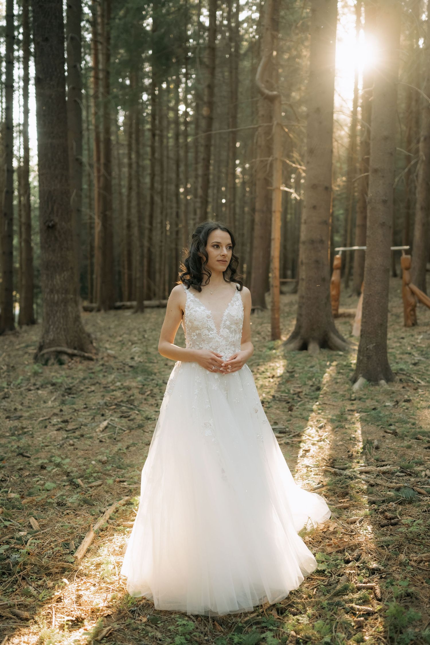 Bride in white lace A-line wedding gown standing in sunlit pine forest, golden hour light streaming through tall trees.