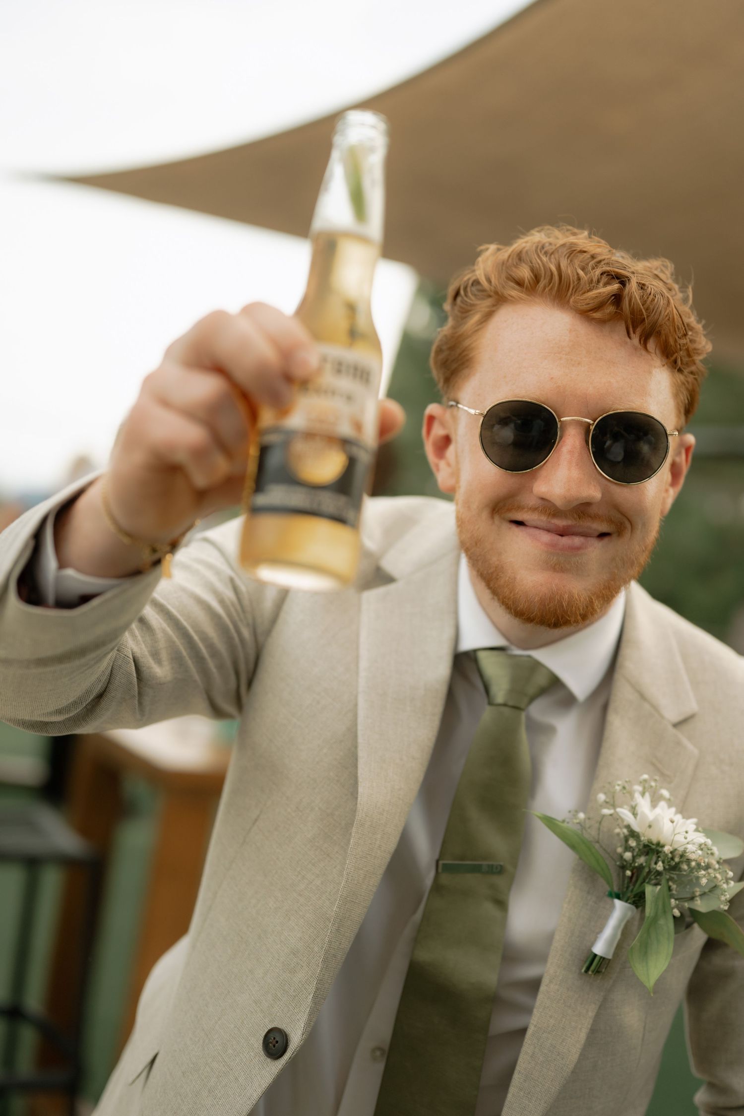 Smiling groom in beige suit and sunglasses raising a Corona beer bottle, with floral boutonniere at outdoor wedding.