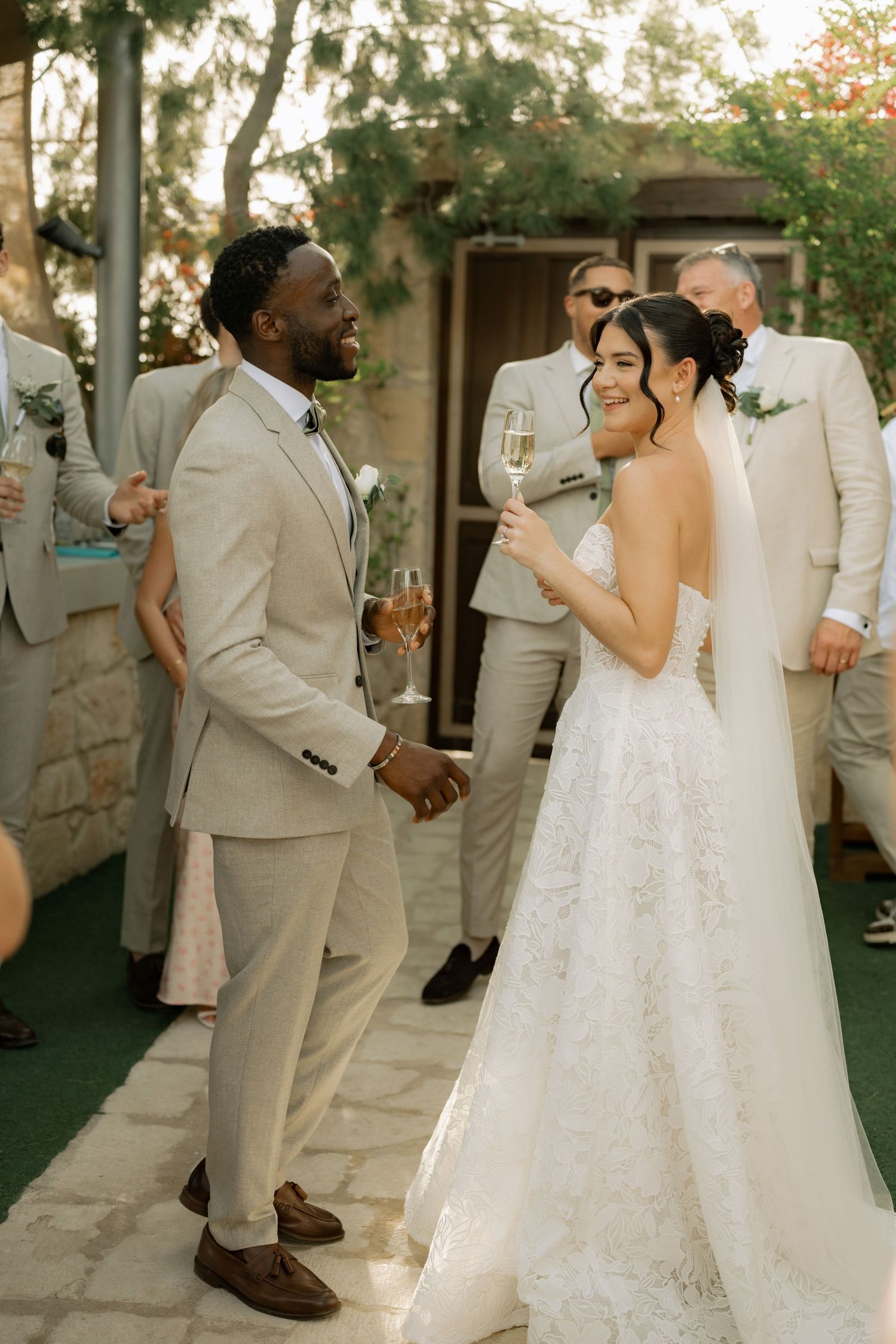 Bride in white lace gown and groom in beige suit toast champagne at outdoor wedding reception.