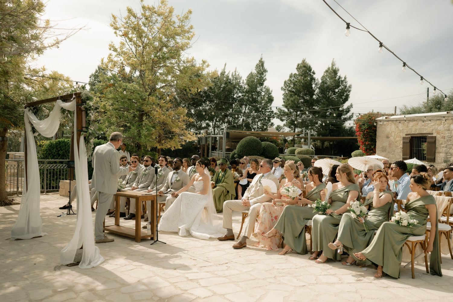 Outdoor wedding ceremony with guests seated in rows, bridesmaids in sage green dresses, under string lights and trees.