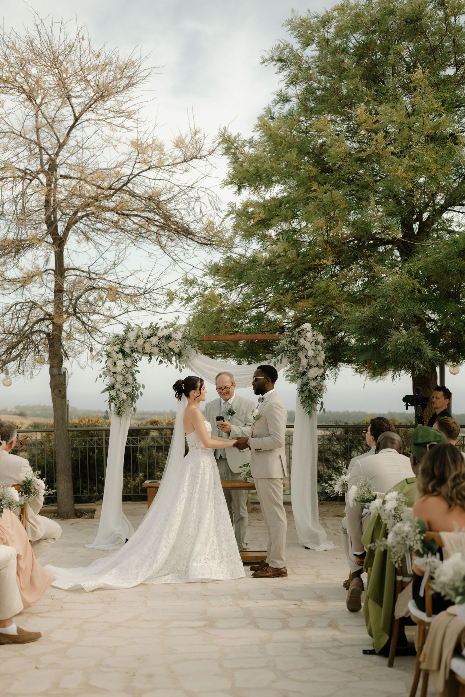 Outdoor wedding ceremony with bride in white gown and groom in tan suit at floral arch, guests seated on patio with scenic views.
