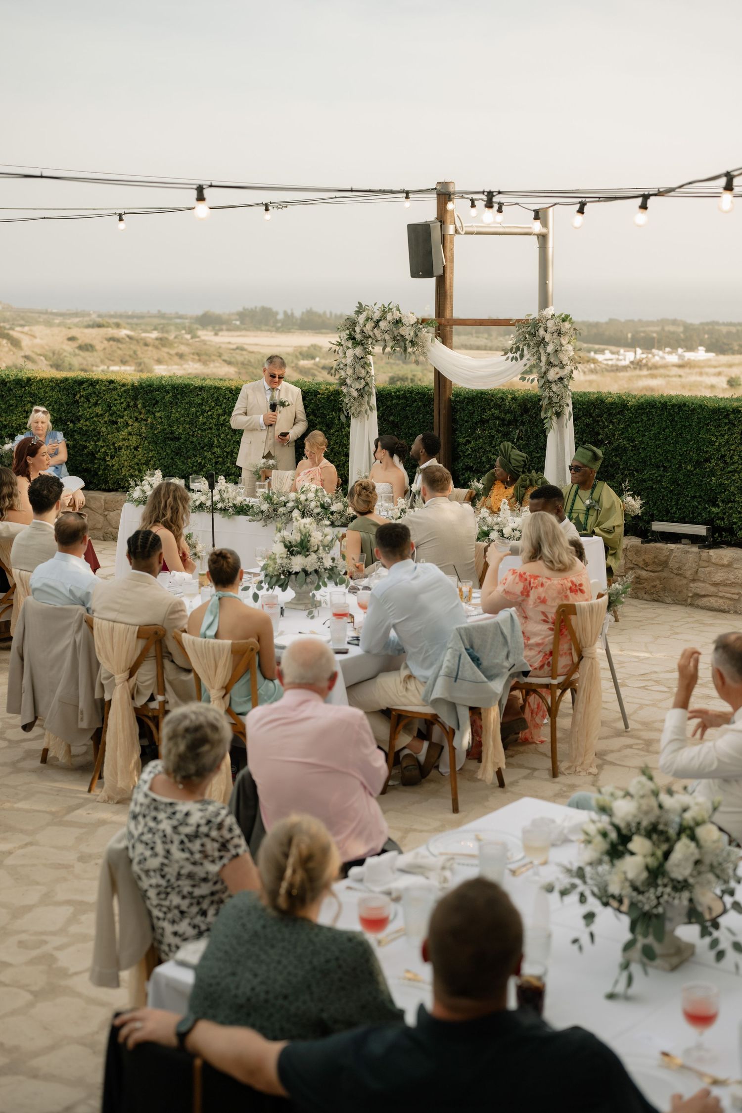 Outdoor wedding reception on a terrace with guests seated at tables, floral arch, string lights, and scenic landscape views.