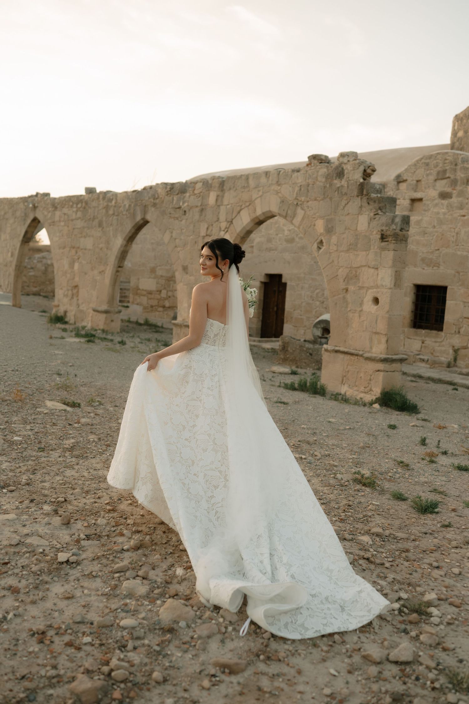 Bride in white lace gown and veil poses before ancient stone arches ruins in arid landscape.