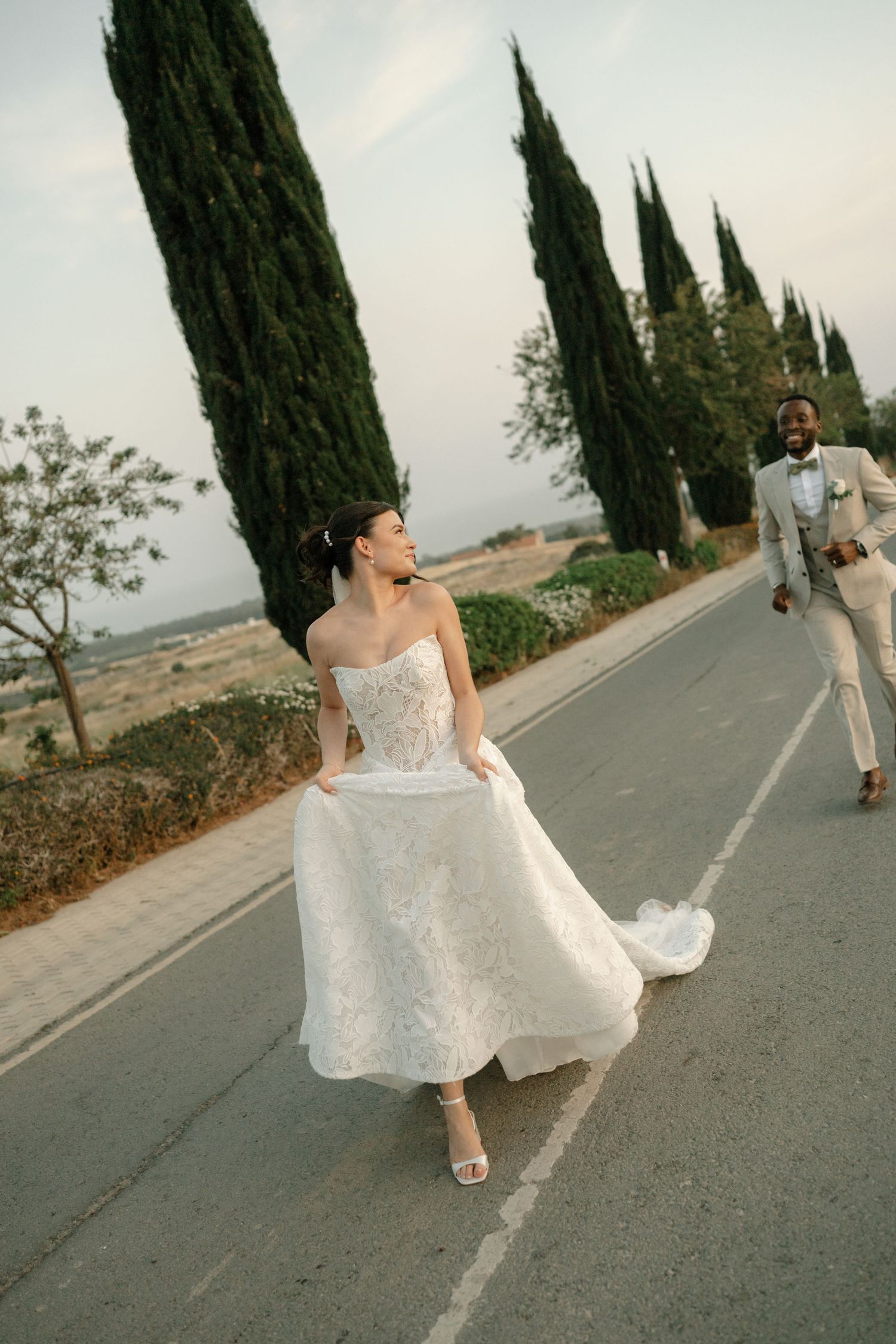 Bride in white strapless gown walks down road lined with cypress trees as groom follows in beige suit.