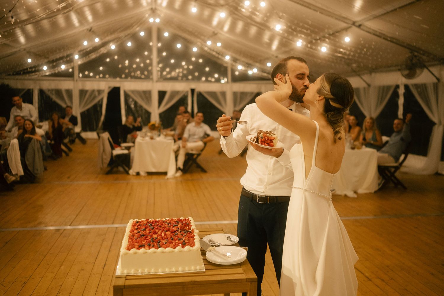 Wedding guests dancing together on a wooden dance floor under string lights in a rustic reception venue.