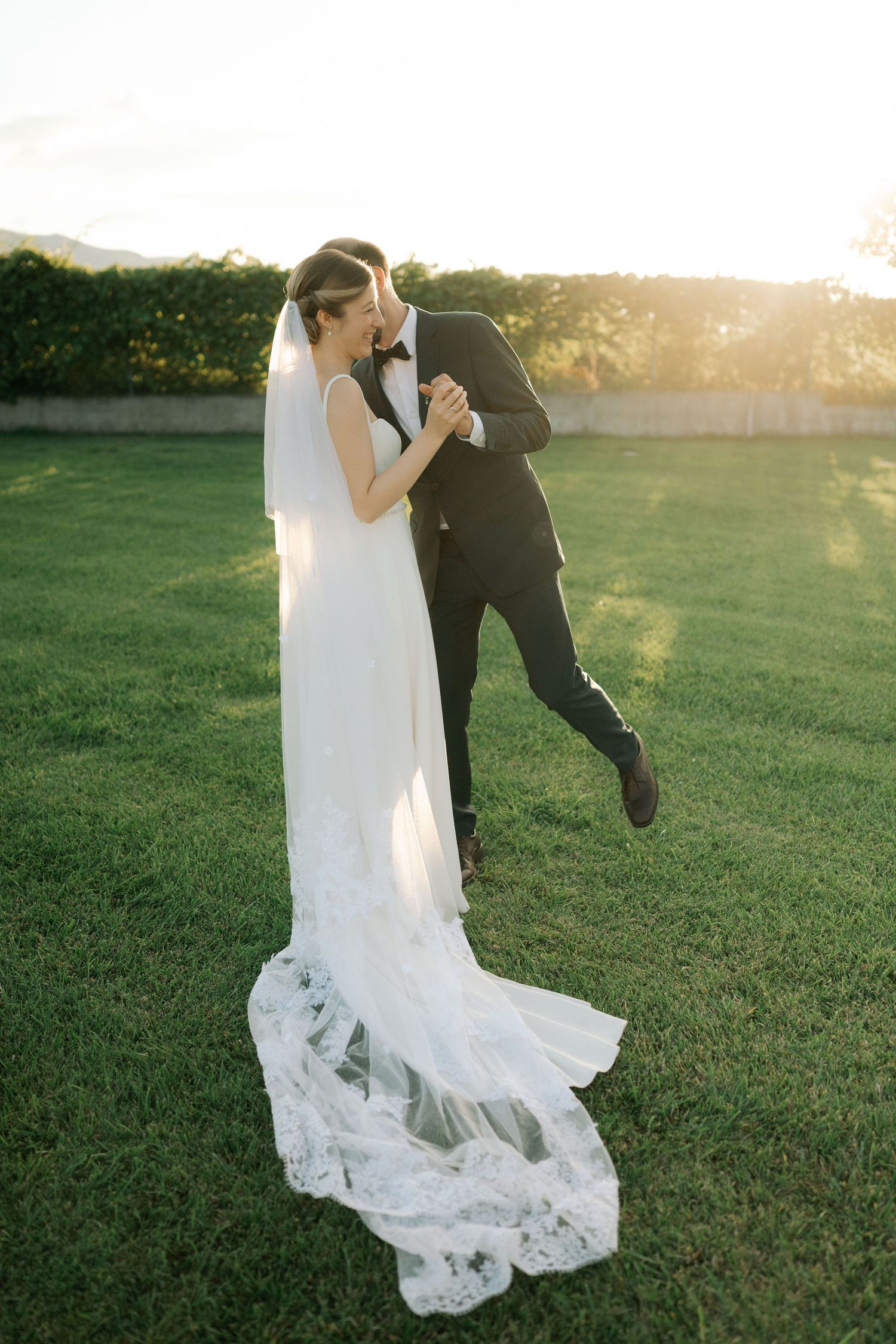 A couple shares a romantic dance in a grassy field at sunset, the bride's lace dress flowing gracefully in the breeze.