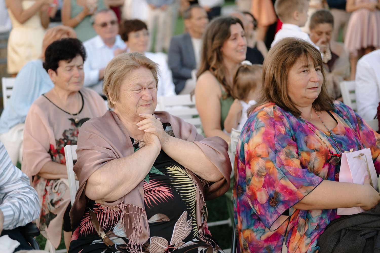 A large group of people sit in an audience at an outdoor event wearing colorful summer attire.