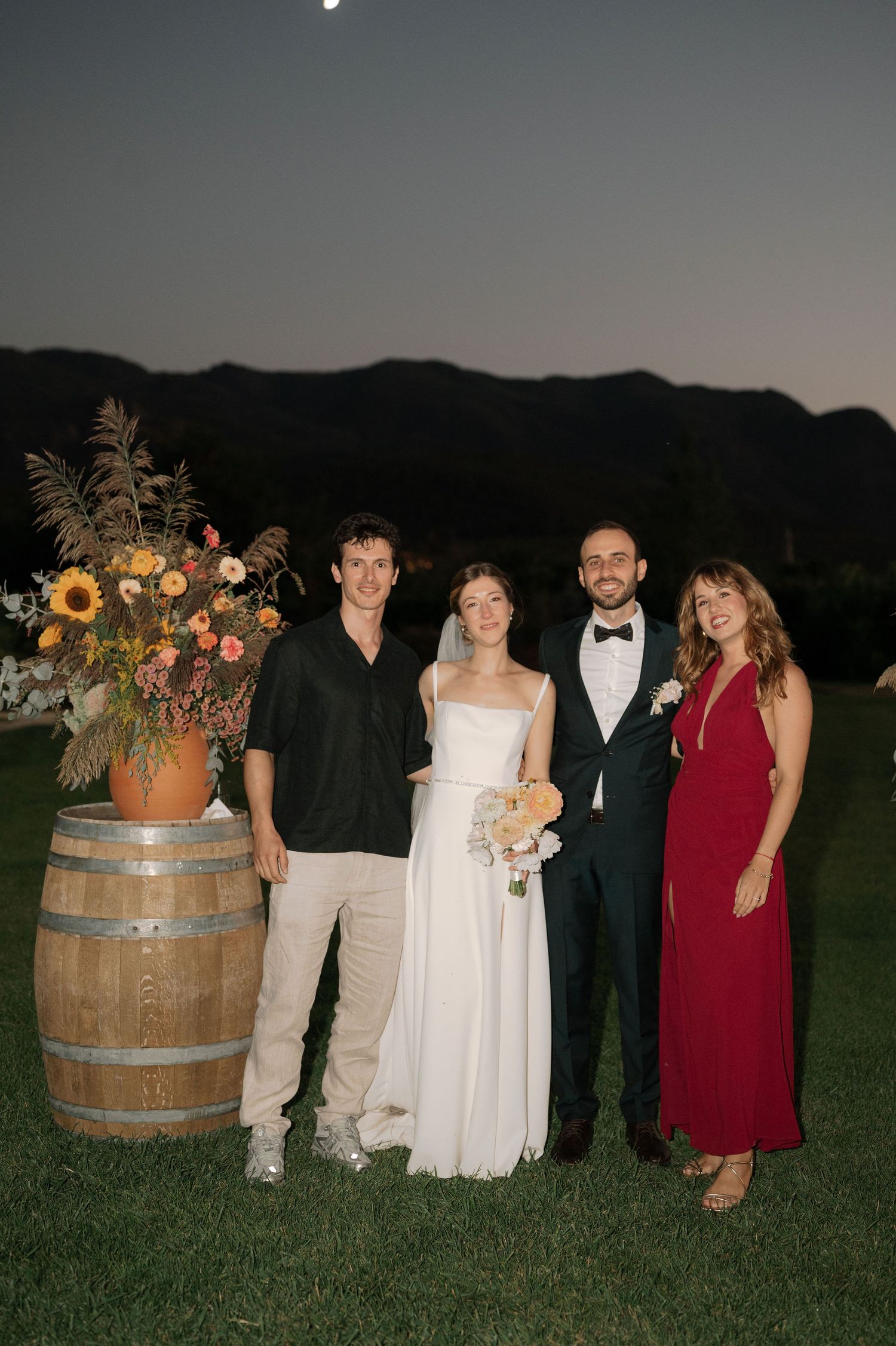 Wedding party poses at sunset with rustic wine barrel and floral arrangements in outdoor mountain setting.