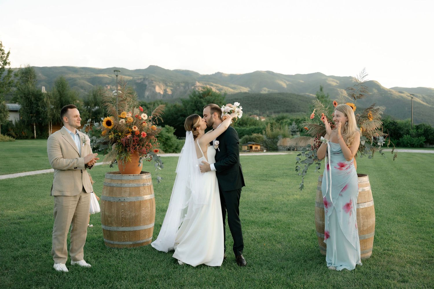 Wedding party gathers around rustic wine barrels at outdoor mountain venue during sunset celebration.