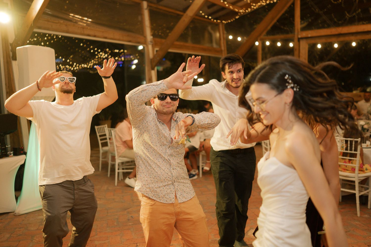 Wedding guests dance in a line formation under string lights at an outdoor evening reception.