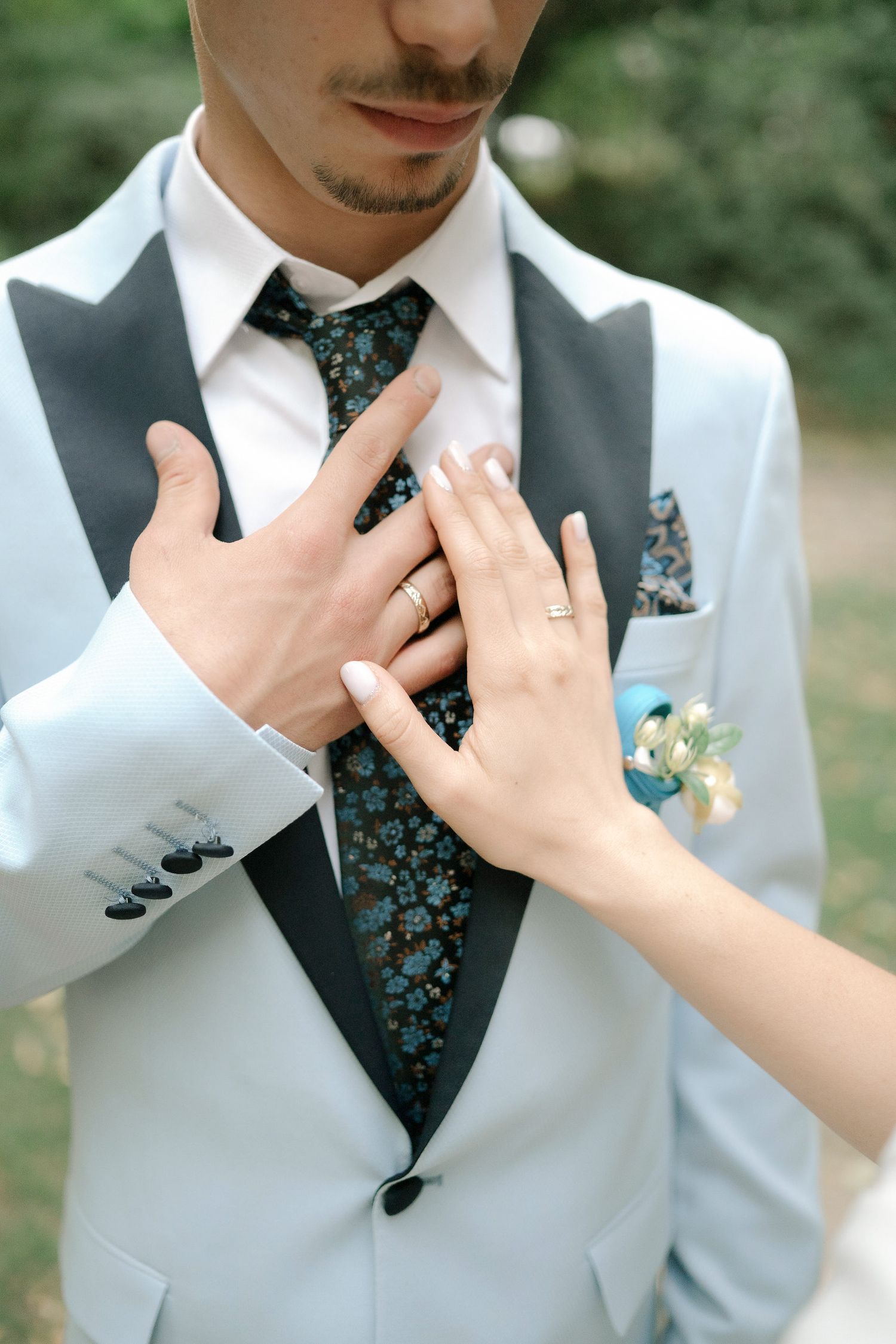 Close-up sequence of hands adjusting a floral patterned tie against a light gray vest at an outdoor wedding ceremony.