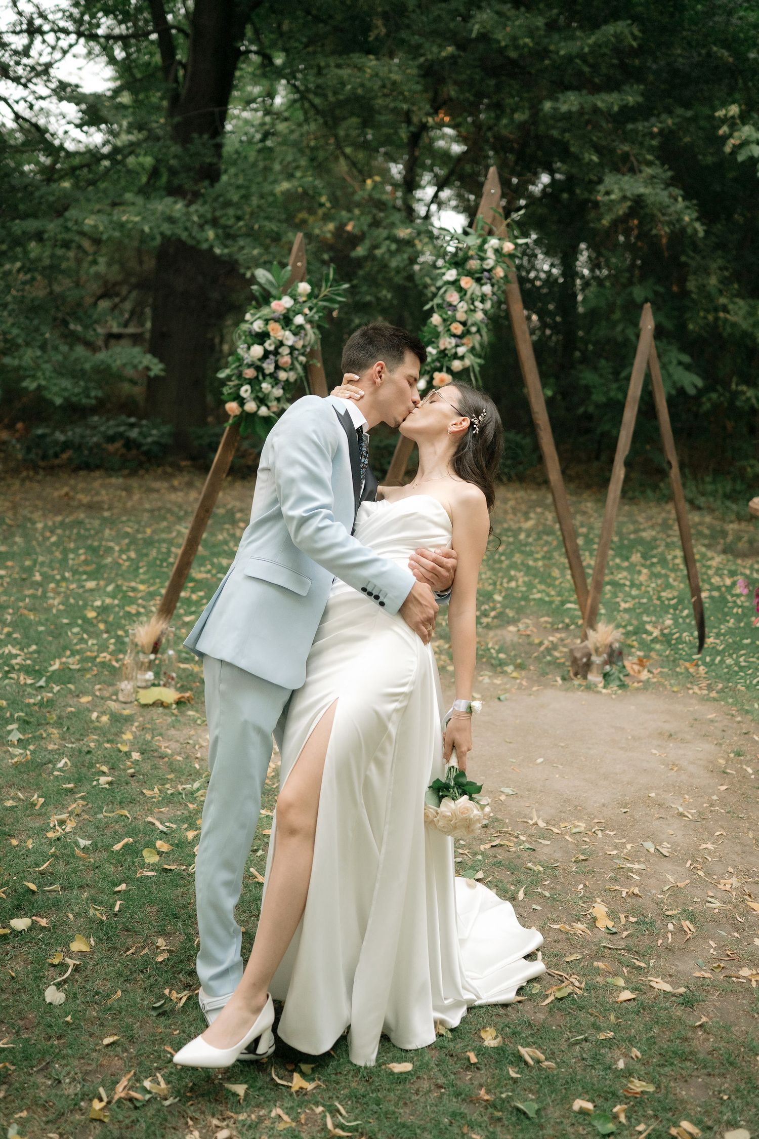 A romantic wedding couple shares intimate moments in a wooded outdoor setting with fallen leaves on the ground.