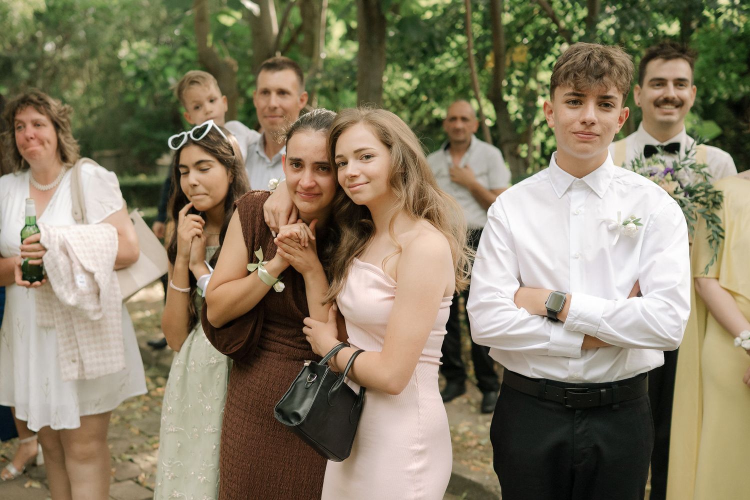 Wedding guests in formal attire stand together outdoors in a natural setting with lush greenery in the background.