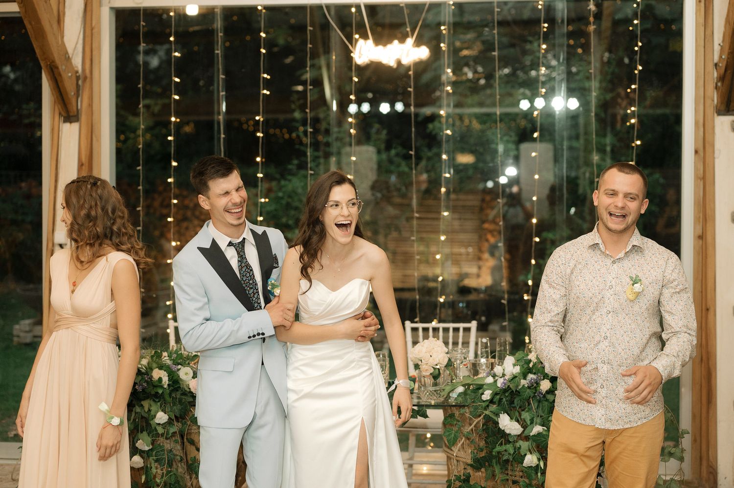 A wedding party celebrates and dances together in a glass-walled venue decorated with twinkling lights.