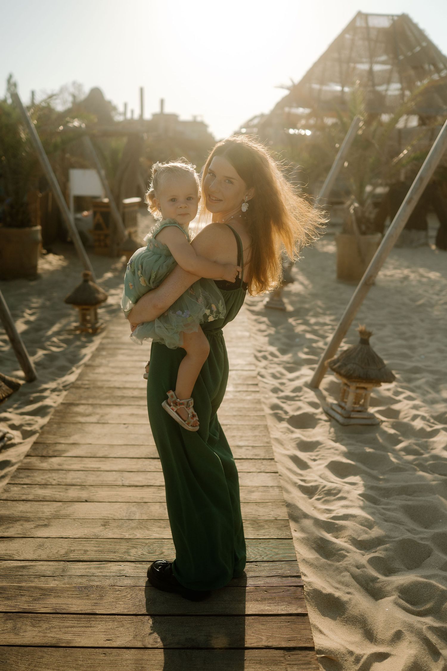 A couple embraces on a wooden boardwalk at sunset with sandy beach and silhouettes in the background.
