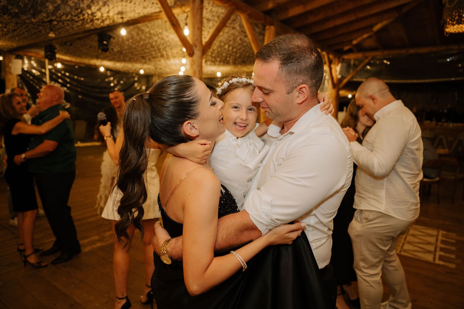 Couples dance together on a rustic wooden dance floor under warm glowing lights at an evening celebration.