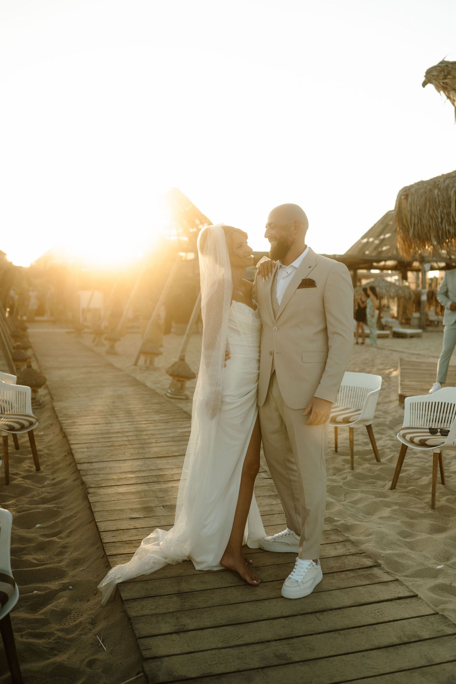 A couple in white wedding attire shares a romantic moment on a wooden pier at sunset.