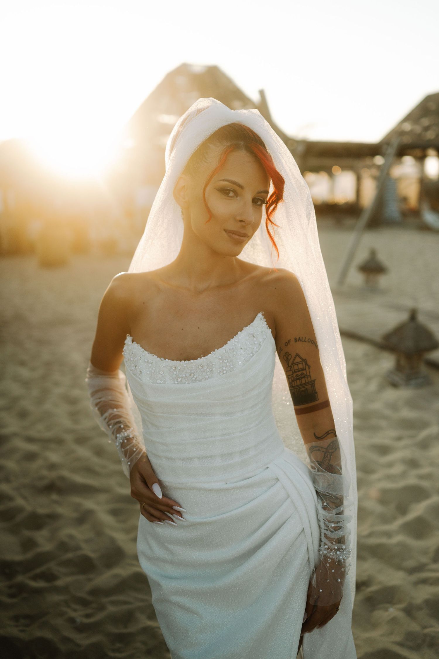 A person in a white wedding dress poses on a sandy beach at sunset with visible arm tattoos.