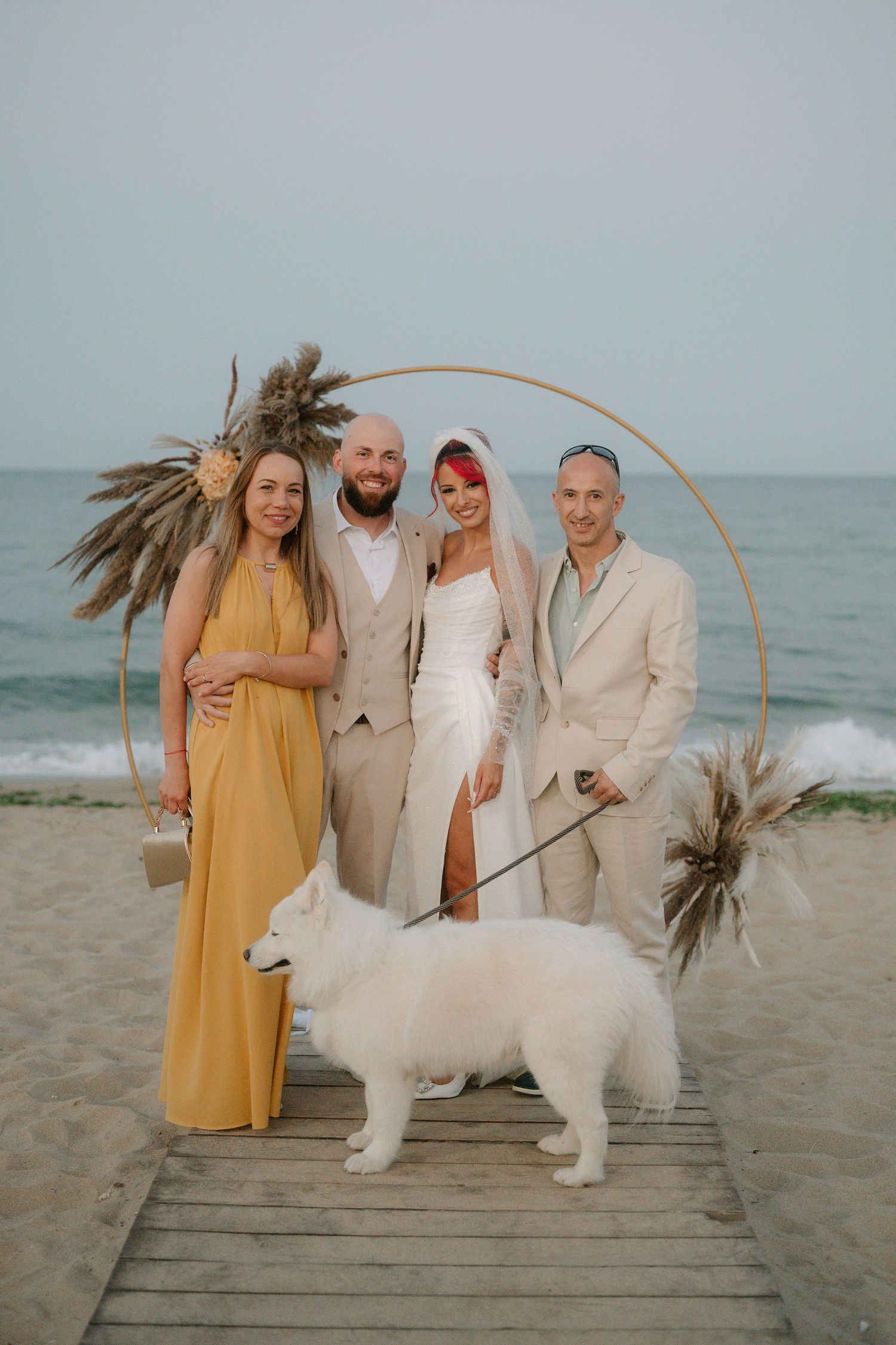 A group poses with a white standard poodle on a wooden boardwalk at the beach during sunset.