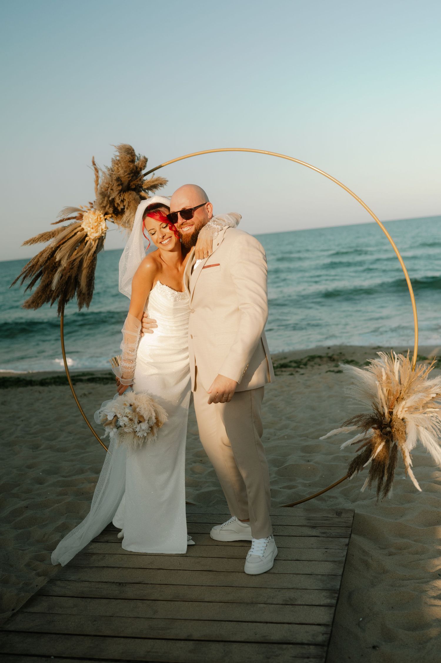 Couple in white attire sharing romantic moments on a wooden boardwalk by the beach at sunset.
