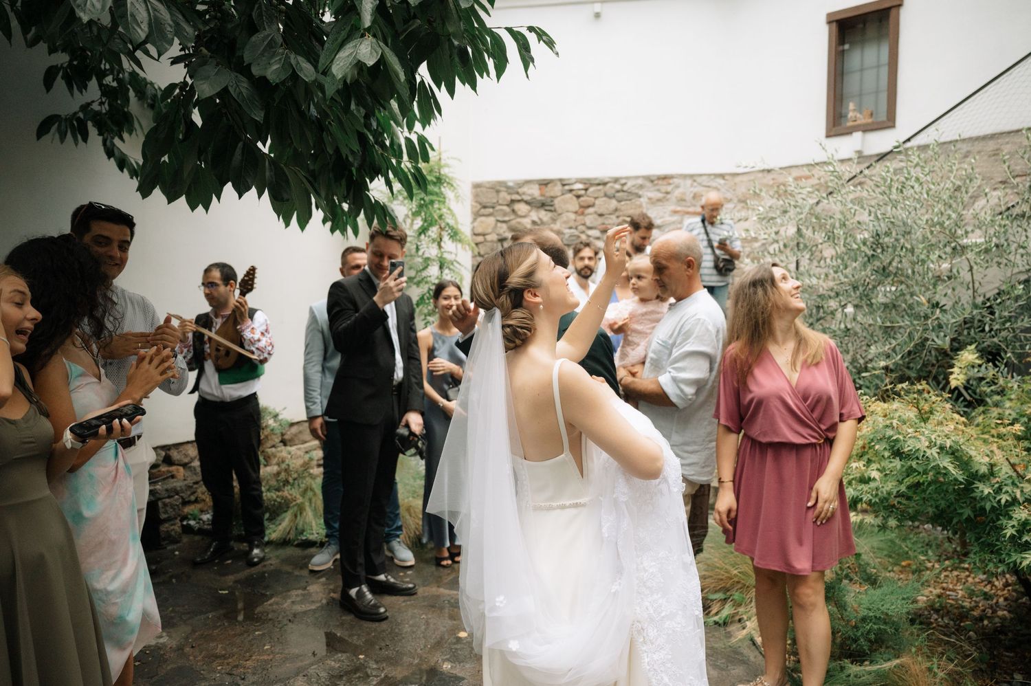 Outdoor wedding celebration with guests gathered among trees and greenery.
