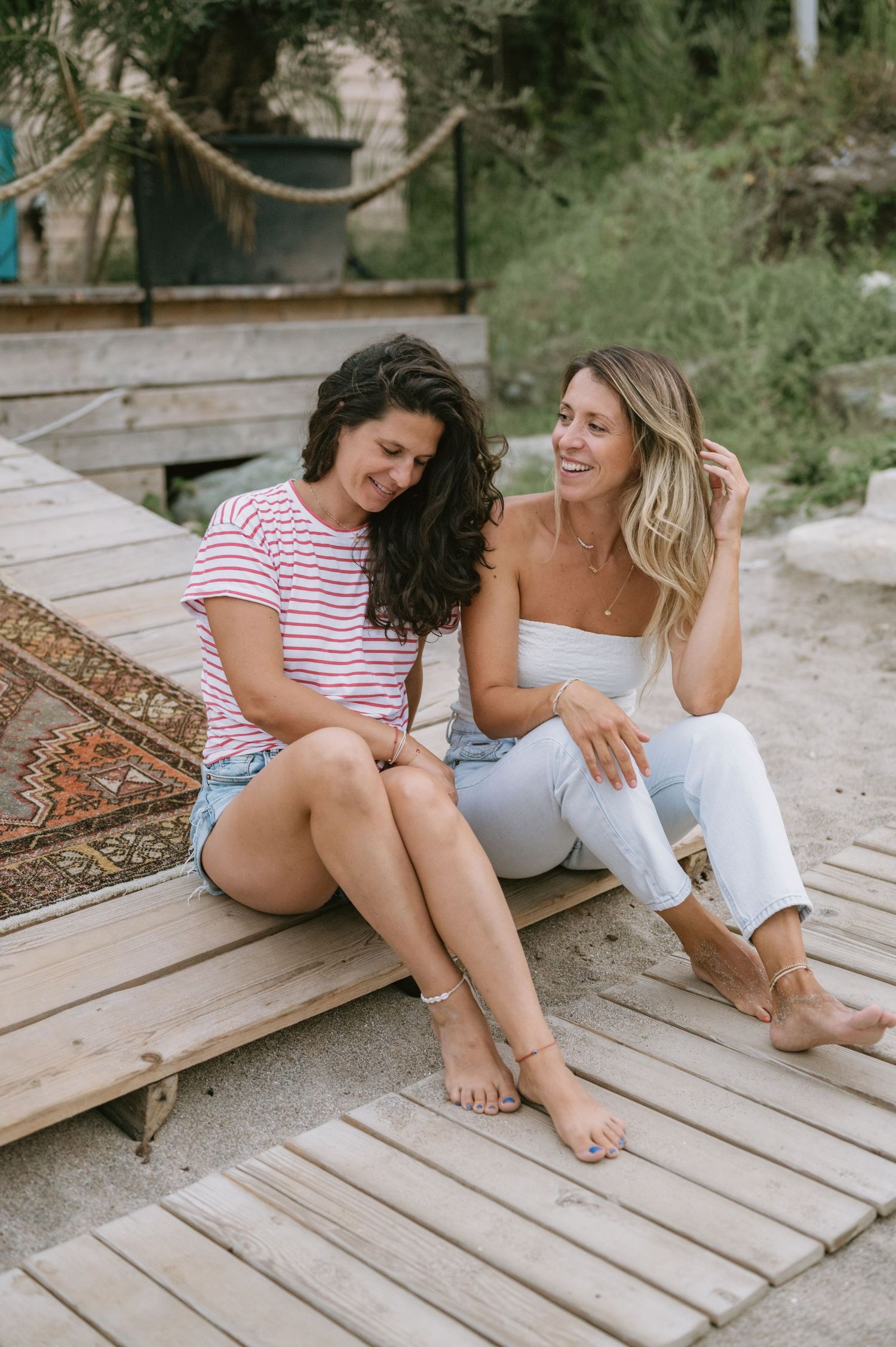 Two women laughing and sitting together on wooden beach steps, one in striped tee and denim shorts, other in white outfit.