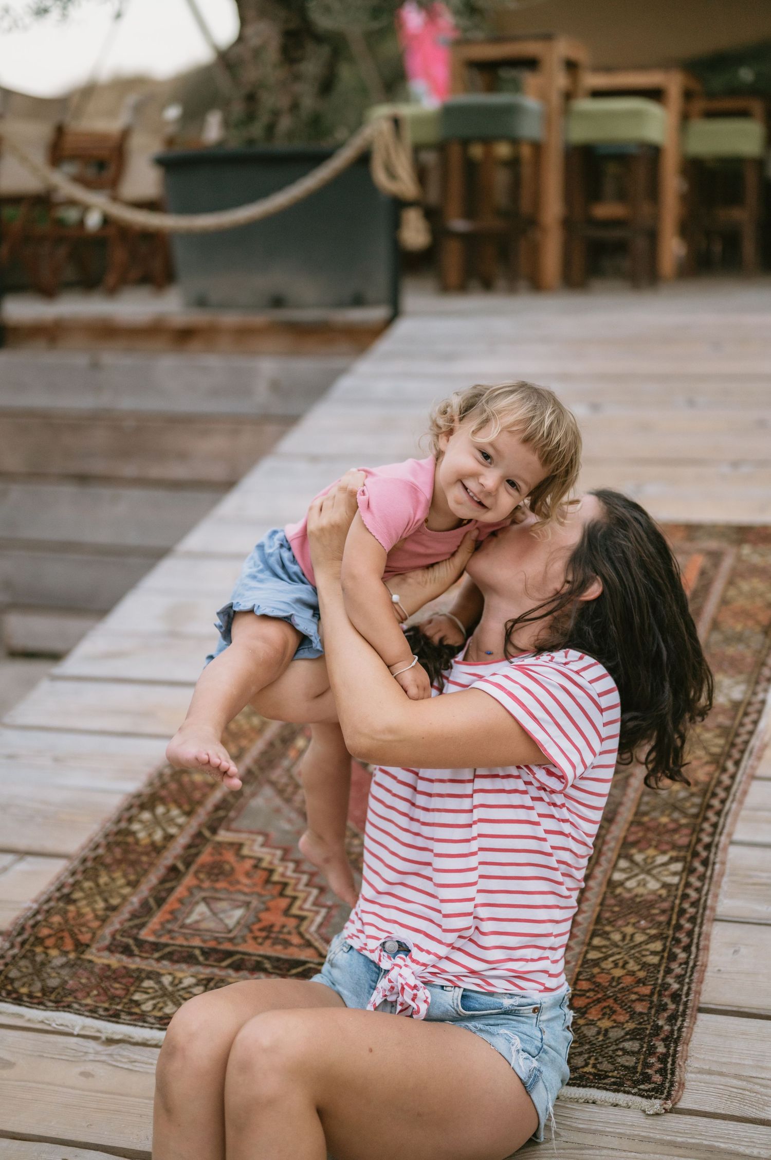 A woman in a striped shirt lifts a smiling toddler on a wooden deck with a colorful rug.