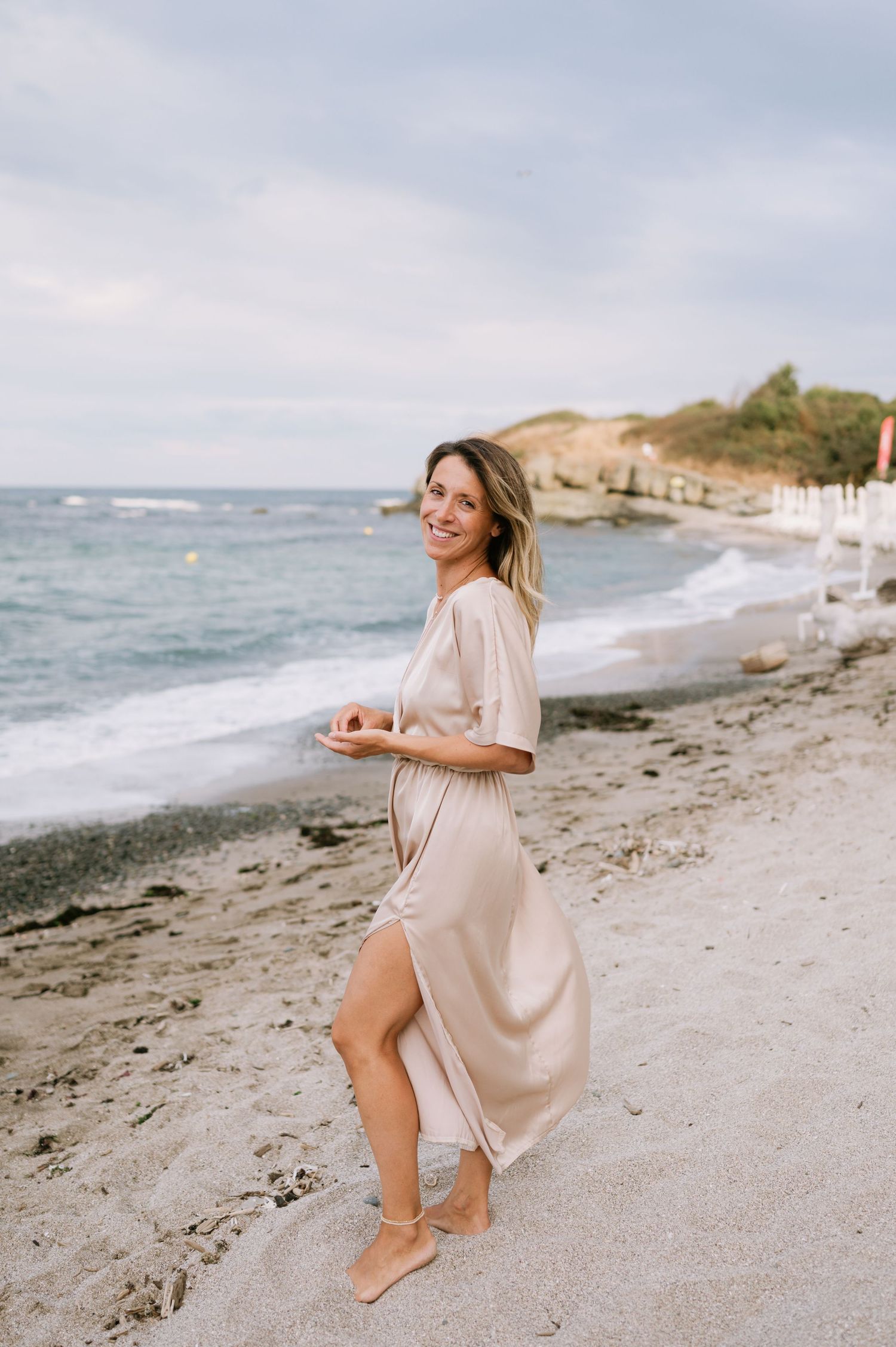 Smiling woman in beige satin dress walking on sandy beach with ocean waves and rocky coastline in background.