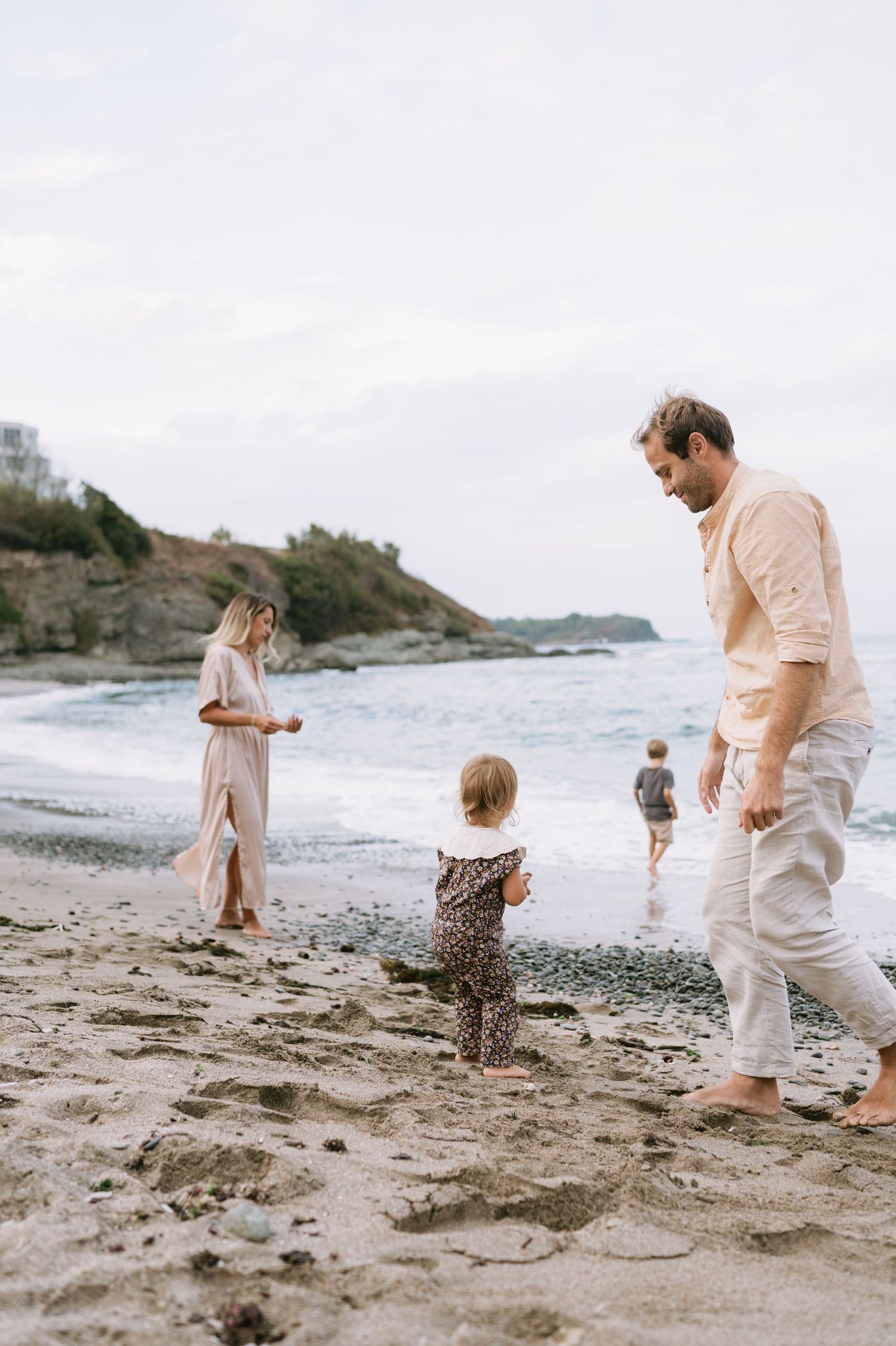 Family of four enjoying a casual beach day, with parents watching their toddler and young child play near the ocean shore.