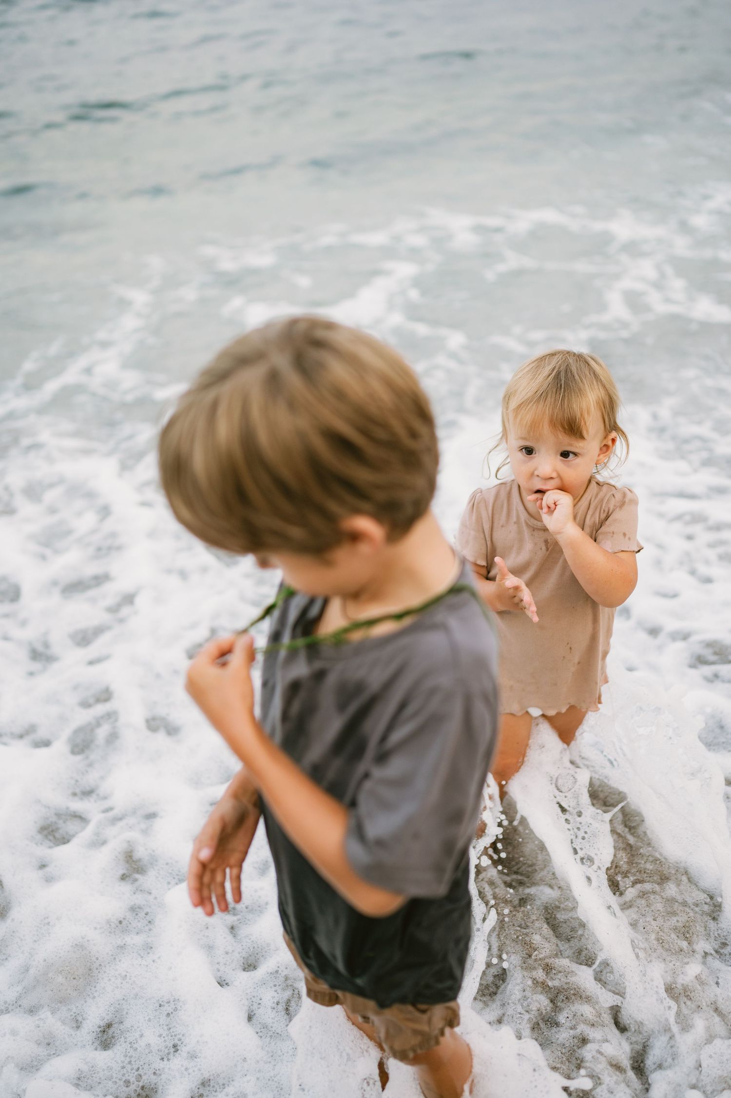 Two young children playing in ocean waves on a sandy beach, with sea foam swirling around their feet.